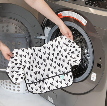 A person puts the Bumkins Sleeved Bib: Hearts into a front-loading washing machine.