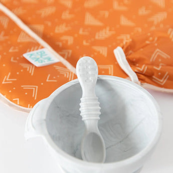 Close-up of a white baby spoon and bowl on a white background, with Bumkins Sleeved Bib: Grounded blurred behind.