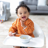 A smiling toddler in Bumkins Sleeved Bib: Grounded sits in a highchair with a spoon and bowl in a bright living room.