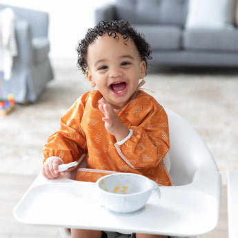 A smiling toddler in Bumkins Sleeved Bib: Grounded sits in a highchair with a spoon and bowl in a bright living room.