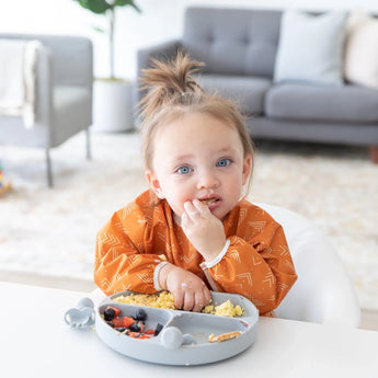 A toddler in a cozy room enjoys baby-led weaning from a gray divided plate, wearing Bumkins Sleeved Bib: Grounded at a white table.