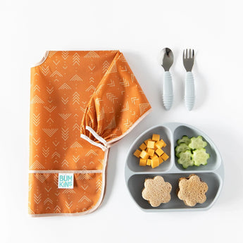Bumkins Sleeved Bib: Grounded lays folded on a white background next to a gray plate with cheese, cucumbers, and sandwiches, and utensils.