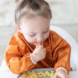 A child wearing a Bumkins Sleeved Bib: Grounded enjoys baby-led weaning by eating with their hands from a gray plate.