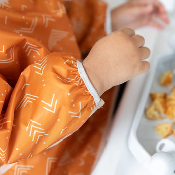 Close-up of a child in a Bumkins Sleeved Bib: Grounded seated at a table, enjoying a baby-led weaning from a white tray.