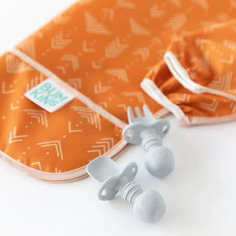 Close-up of gray baby utensils next to a folded Bumkins Sleeved Bib: Grounded on a white background.