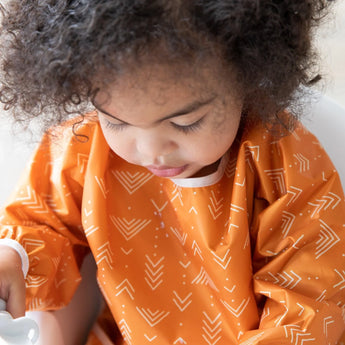 A curly-haired child in a Bumkins Sleeved Bib: Grounded looks down, ready for baby-led weaning.