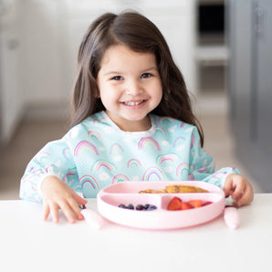 A happy girl in a Bumkins Sleeved Bib: Rainbows enjoys a meal from a pink plate at a white table.
