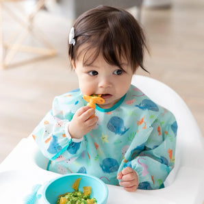 A toddler in a Bumkins Sleeved Bib: Ocean Life sits in a high chair, chewing an orange spoon with a blue bowl on the tray.
