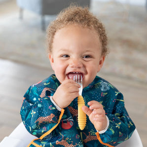 A happy toddler in a Bumkins Sleeved Bib: Jungle holds an orange fork near their mouth.