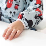 A childs hand on a white surface in a Bumkins sleeved bib with red and black Mickey Mouse designs on a light gray shirt.