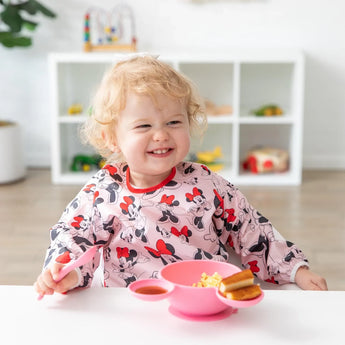 A toddler in a Bumkins Minnie Mouse sleeved bib smiles, enjoying baby-led weaning from a pink bowl with toys on white shelves behind.