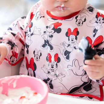 A child joyfully explores yogurt with a spoon in a Bumkins Waterproof Minnie Mouse Sleeved Bib, face and pink bowl decorated.