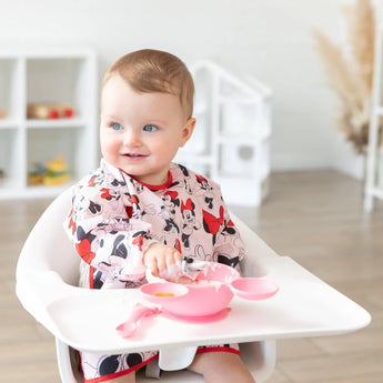 A baby in a Bumkins Sleeved Bib: Minnie Mouse holds a spoon at a tray with pink dishes, set against a neutral background.