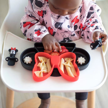 A child in a Bumkins Sleeved Bib: Minnie Mouse sits in a high chair, enjoying pasta and strawberries from a bow-shaped plate.