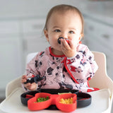 A toddler in a high chair enjoys vibrant food with a spoon, wearing a colorful Bumkins Sleeved Bib: Minnie Mouse.