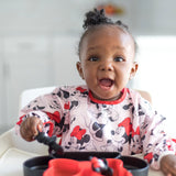 A joyful baby uses a Bumkins Sleeved Bib: Minnie Mouse, holding a spoon with a divided plate on a highchair.