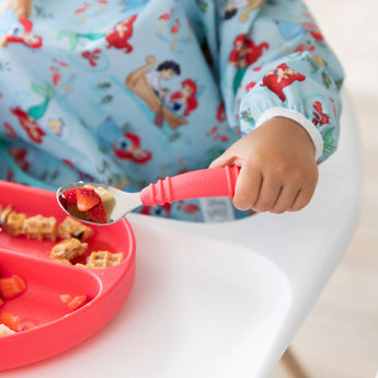 Close-up of a child in Bumkins Sleeved Bib: Ariel holding a spoon with fruit over a red plate with waffle pieces on a high chair tray.