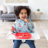 A child with curly hair in a high chair, wearing Bumkins Sleeved Bib: Ariel, smiles while holding a spoon next to a red plate on the tray.