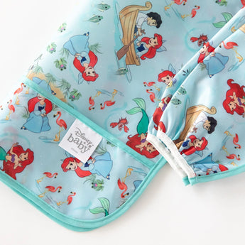 Close-up of Bumkins Sleeved Bib: Ariel folded on a white background, with focus on the front pocket and elastic cuff on the sleeves. 
