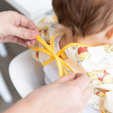 Close-up of adult hands tying the ribbon closure of a Bumkins Sleeved Bib: Winnie Loves Hunny around a child's neck.