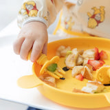 Close-up of a child wearing a Bumkins Sleeved Bib: Winnie Loves Hunny, their hand using a fork to eat diced fruits from a yellow plate.