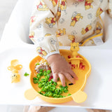 A baby in a high chair practicing self-feeding with peas on a bear-shaped plate, wearing a Bumkins Sleeved Bib: Winnie Loves Hunny.