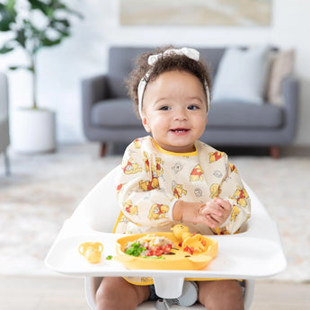 A toddler in a high chair claps hands, wearing a Bumkins Sleeved Bib: Winnie Loves Hunny, with food on a plate in a bright room.