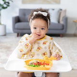 A toddler in a Bumkins Sleeved Bib: Winnie Loves Hunny sits in a high chair, eating from a yellow plate in a cozy living room.