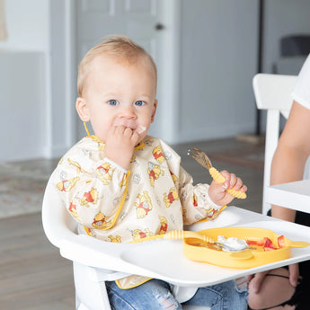 A baby in a high chair, wearing a Bumkins Sleeved Bib: Winnie Loves Hunny, enjoys baby-led weaning with a yellow fork and plate.