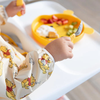 Close-up of a child in a high chair wearing a Bumkins Sleeved Bib: Winnie Loves Hunny, holding a fork and spoon over a yellow plate.