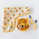 Bumkins Sleeved Bib: Winnie Loves Hunny lays folded next to a yellow bear-shaped plate of food and yellow utensils on a white background.
