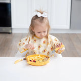 A toddler in a Bumkins Sleeved Bib: Winnie Loves Hunny uses a fork to eat from a yellow plate, exploring baby-led weaning at a white table.