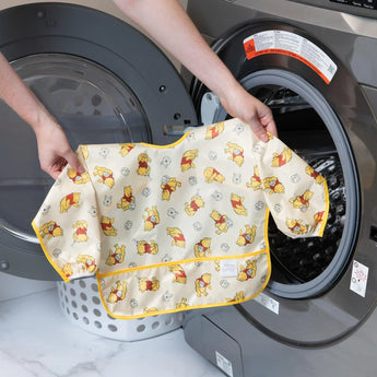 A person holds Bumkins Sleeved Bib: Winnie Loves Hunny in front of an open front-loading washing machine.