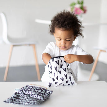 Toddler opens a Bumkins Reusable Snack Bag in Hearts design at a table, next to another bag from the Large 2-Pack: XOXO & Hearts.