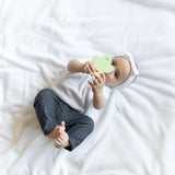 A baby in a white shirt and gray pants lies on a white blanket, holding a Bumkins Silicone Teether: Cactus, wearing a white headband.