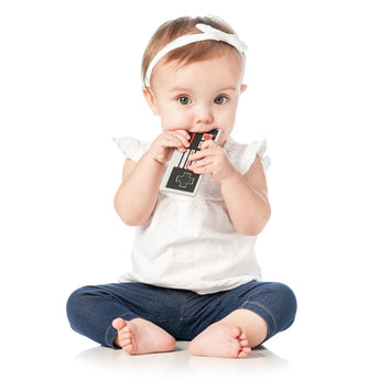 A baby sits on the floor, chewing on a Bumkins Silicone Teether: NES™ Controller, for gum relief.