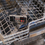 A Silicone Teether: NES™ Controller by Bumkins sits in the dishwasher, ready to soothe babys gums with nostalgic charm.