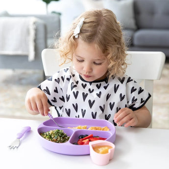 A curly-haired child in a Bumkins Hearts bib eats from a purple divided plate, wearing a shirt with black hearts.
