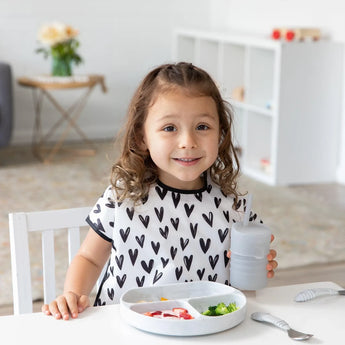 A curly-haired girl sits at the table in a stain-resistant Bumkins Junior Bib: Hearts, holding a cup and eyeing her plate of food.