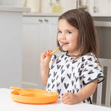 A girl in a Bumkins Junior Bib: Hearts sits at the table, brushing her teeth with an orange toothbrush.