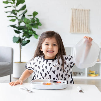 A child wearing Bumkins Junior Bib: Hearts lifts a tray lid, with a plant in the background. The bib fits snugly and is stain-resistant.