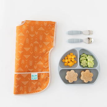 Bumkins Junior Bib: Grounded is folded on a white background next to a gray plate with cheese, cucumbers, and sandwiches, and gray utensils.