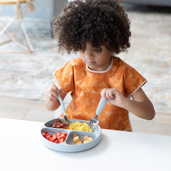 A child in a Bumkins Junior Bib: Grounded enjoys breakfast from a gray divided plate at a white table.