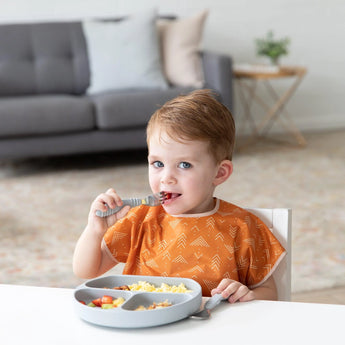 A toddler wearing a Bumkins Junior Bib: Grounded munches from a gray divided plate. A comfy couch and side table are in the background.