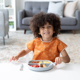 A curly-haired child beams in a Bumkins Junior Bib: Grounded, sitting at a table with food. A cozy sofa and shelves fill the background.