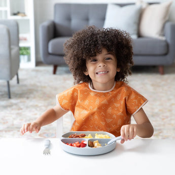 A curly-haired child beams in a Bumkins Junior Bib: Grounded, sitting at a table with food. A cozy sofa and shelves fill the background.