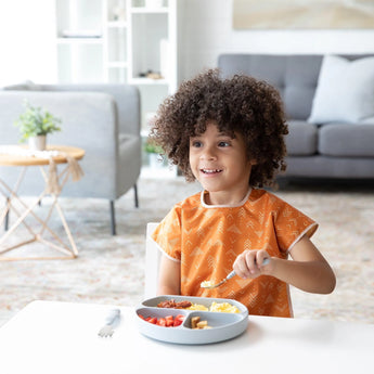 A child with curly hair enjoys a meal from a gray divided plate, wearing a Bumkins Junior Bib: Grounded in a bright living room.