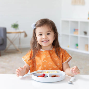 A smiling girl with long hair and a flower clip wears a Bumkins Junior Bib: Grounded while seated at a table with a divided plate of food.