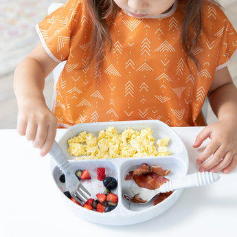 A child eats eggs and fruit from a gray plate at a white table, wearing a Bumkins Junior Bib: Grounded.