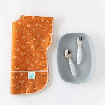Bumkins Junior Bib: Grounded lies folded next to a grey plate with a child-sized fork and spoon on a white background.